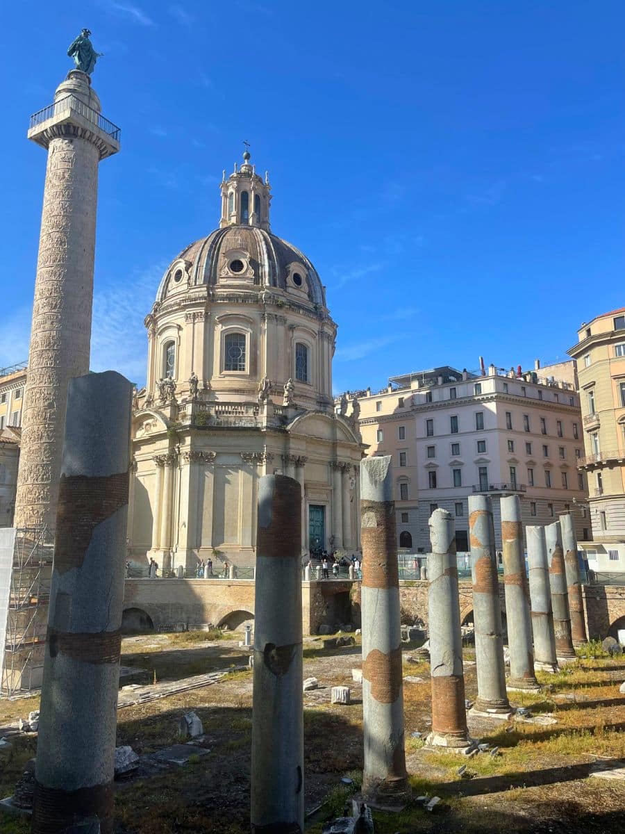 A historic site in Rome, Italy, featuring Trajan's Column, an intricately carved monument topped with a statue, and the grand dome of the Church of the Most Holy Name of Mary. In the foreground, ancient ruins with tall, weathered stone columns stand, showcasing remnants of Rome's past. The bright blue sky contrasts beautifully with the earthy tones of the ruins and the Baroque-style church. A few visitors explore the site, adding a sense of scale and liveliness to the scene.