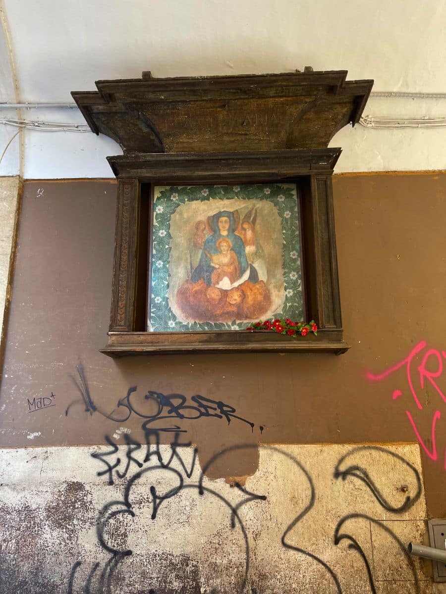 A religious painting of the Virgin Mary and Child with angels, housed in an old wooden shrine mounted on a weathered brown wall in Rome, Italy. The words 'Ave Maria' are inscribed on the wooden frame. Below the shrine, red flowers are placed as an offering. The surrounding wall is covered in graffiti, contrasting the sacred image with modern urban decay.