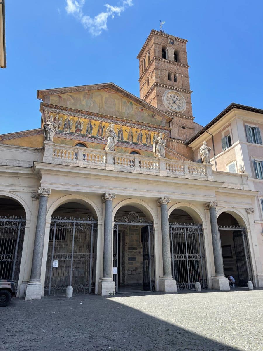 The Basilica of Santa Maria in Trastevere, one of the oldest churches in Rome, Italy. The facade features a beautiful golden mosaic depicting religious figures, with a row of statues on the balcony below. The Romanesque-style bell tower stands tall, adorned with a clock. The entrance is framed by arched porticos with iron gates. The cobblestone piazza in front is partially shaded, with a clear blue sky above