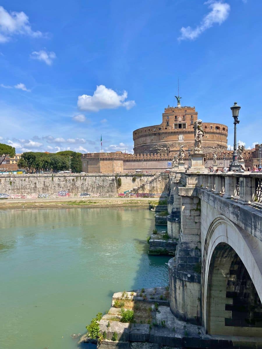 A view of Castel Sant'Angelo in Rome, Italy, as seen from Ponte Sant’Angelo. The cylindrical fortress, originally built as Emperor Hadrian’s mausoleum, stands against a bright blue sky. The bridge, adorned with statues of angels, stretches across the Tiber River, whose green waters reflect the surrounding historic architecture.