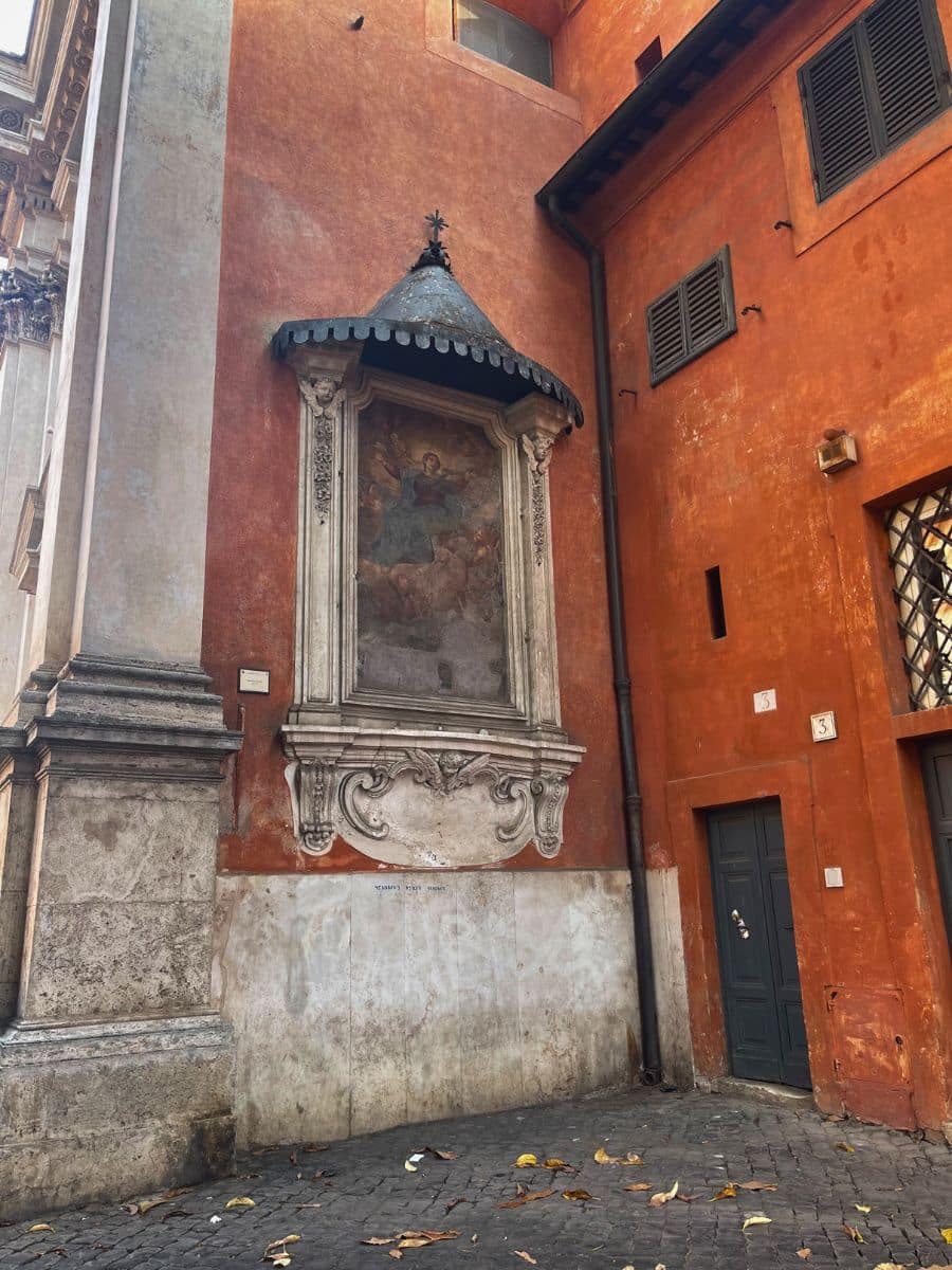 A historic street corner in Rome, Italy, featuring an old fresco painting set within an ornate stone frame on a warm, ochre-colored wall. The fresco is protected by a small, metal canopy with decorative trim. A mix of architectural elements, including shuttered windows, a weathered stone base, and aged plaster, adds character to the scene. Fallen leaves are scattered on the cobblestone street below, enhancing the timeless charm of the location.