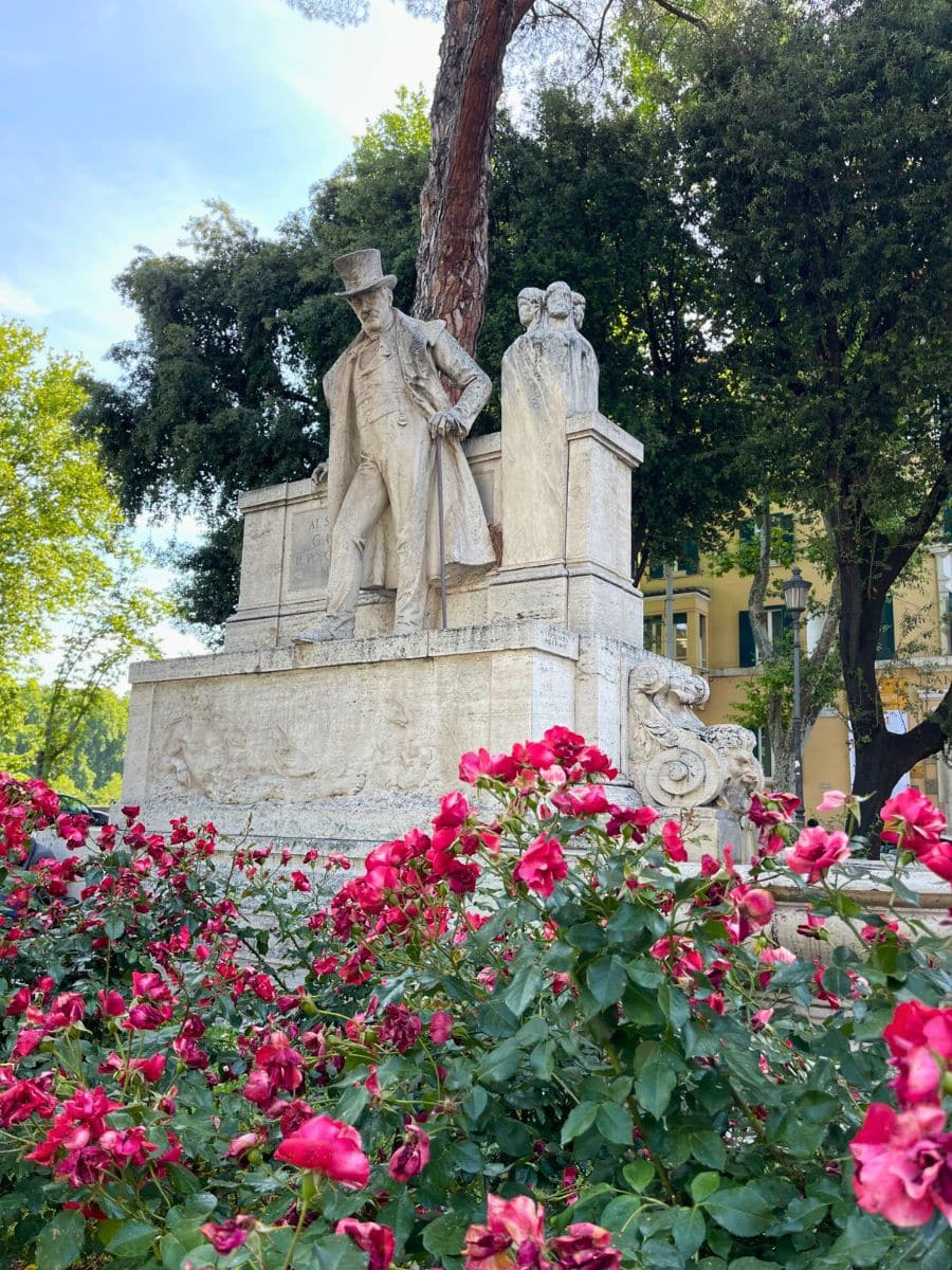 A marble statue in a lush park in Rome, Italy, surrounded by vibrant red roses. The statue depicts a man wearing a top hat and a long coat, leaning forward with a cane, with a solemn expression. Behind him, there are sculpted figures on the monument, adding depth to the composition. A tall tree grows beside the monument, and in the background, a yellow building with green shutters is partially visible through the foliage. The scene is bathed in natural daylight, enhancing the contrast between the stone sculpture and the colorful flowers.