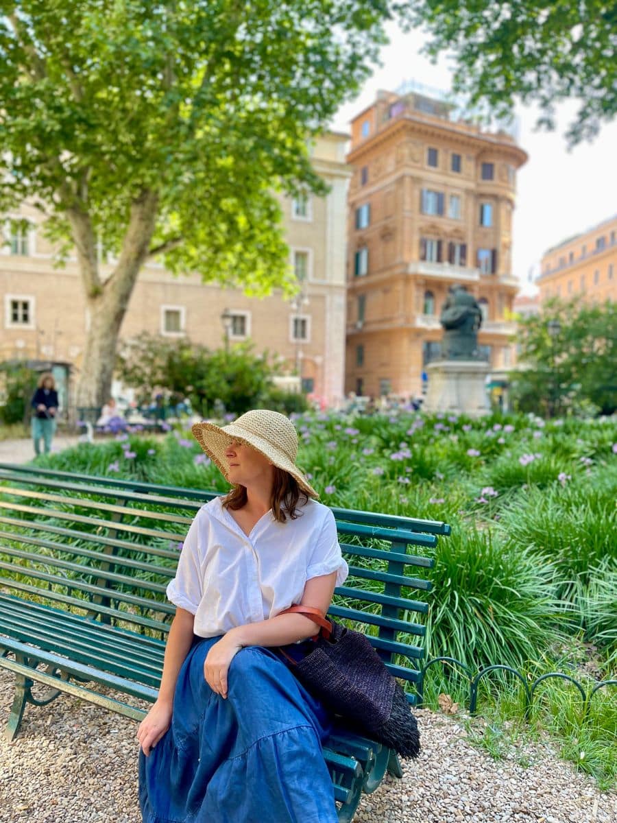 Melissa wearing a straw hat, white blouse, and flowing blue skirt sits on a green park bench in a lush garden in Rome, Italy. She gazes to the side with a relaxed expression, holding a woven black bag with brown straps. The park is filled with greenery, blooming purple flowers, and tall trees that provide shade. In the background, historic buildings with ornate facades and shuttered windows line the scene, along with a statue on a pedestal. A few people are seen strolling through the park, enjoying the tranquil atmosphere under the warm sunlight.
