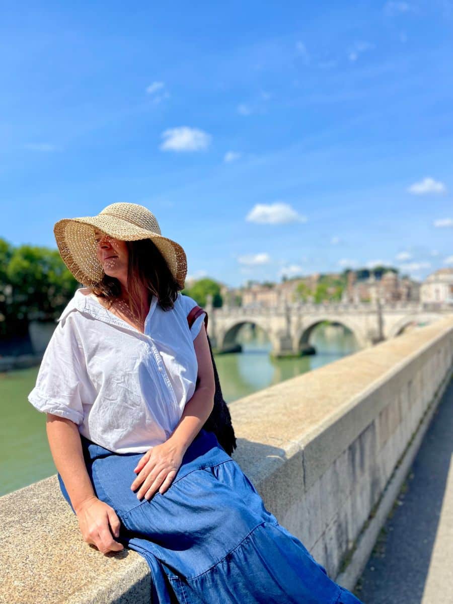 A woman sits on the stone railing of a bridge in Rome, Italy, basking in the warm sunlight. She wears a wide-brimmed straw hat that casts a shadow over her face, a loose white button-up shirt, and a flowing blue skirt. A black tote bag hangs from her shoulder. The background showcases the Tiber River and an old stone bridge with elegant arches, with the cityscape of Rome visible beyond. The sky is bright blue with a few scattered clouds, and the sunlight highlights the serene atmosphere of the moment.