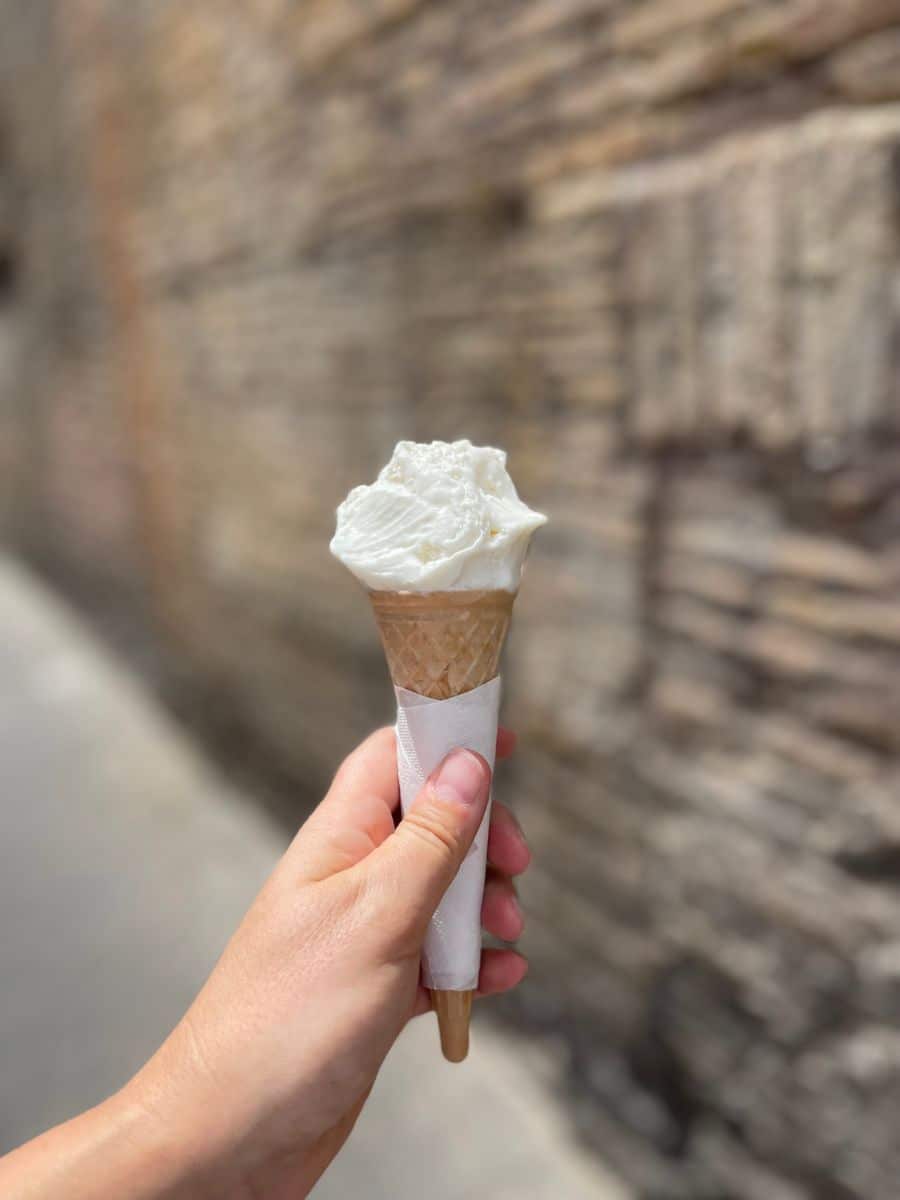 A hand holding a cone of creamy white gelato, wrapped in a napkin, against the backdrop of an old brick wall in Rome, Italy. The gelato appears slightly melting, capturing the essence of enjoying a cool treat on a warm day. The blurred background emphasizes the texture of the aged bricks, adding a sense of history and charm to the moment.