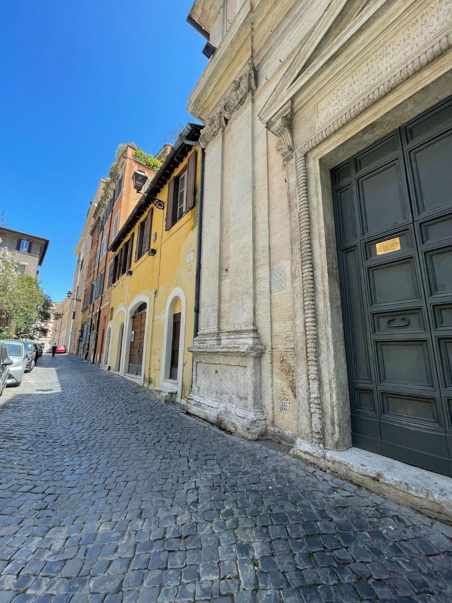 A charming cobblestone street in Rome, Italy, lined with historic buildings. On the right, a grand stone structure with intricate carvings and a dark green wooden door stands tall. On the left, a row of ochre and yellow buildings with wooden shutters and arched doorways exudes old-world charm. Small balconies with greenery add a touch of life to the scene. The bright blue sky and warm sunlight highlight the textures of the stone and brick, creating a picturesque Roman street atmosphere.