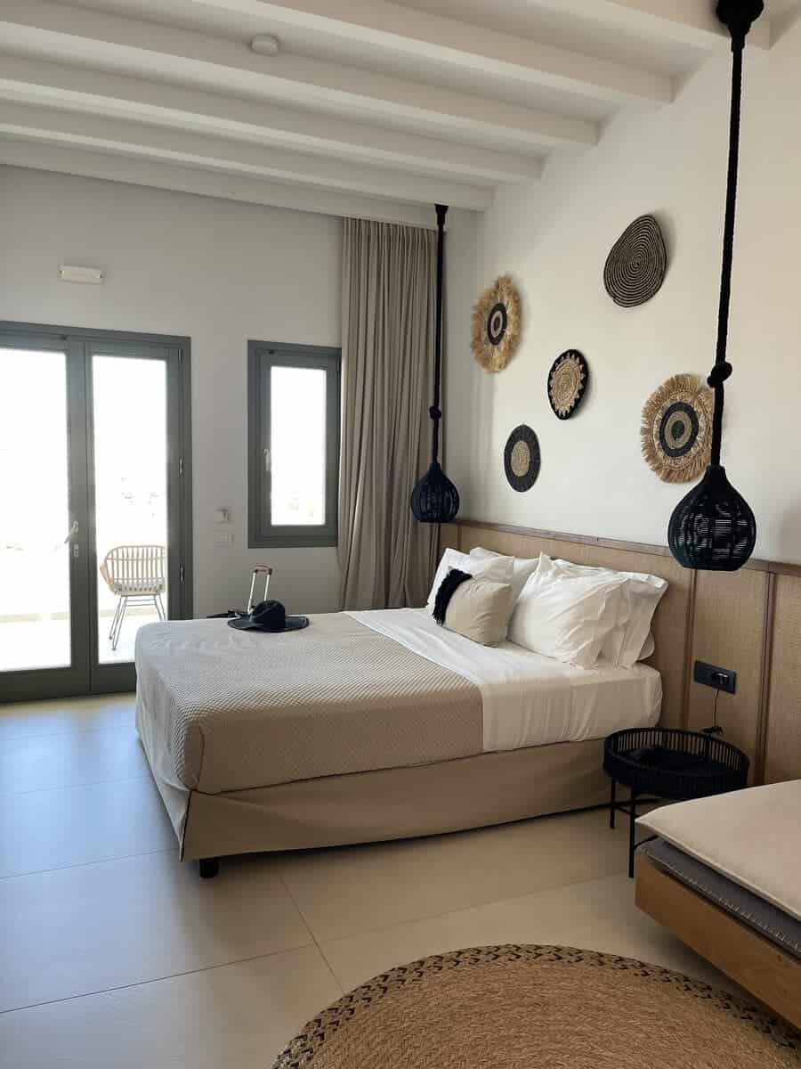 A modern, minimalist bedroom with a white bed, black pendant lights, wicker wall decor, and large windows leading to a balcony. The design is clean and airy.