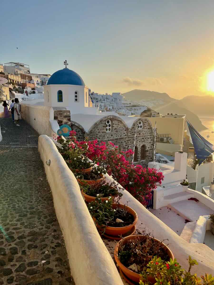 Iconic Santorini scene with a whitewashed walkway, a blue-domed church, and vibrant pink flowers overlooking the Aegean Sea at sunset.