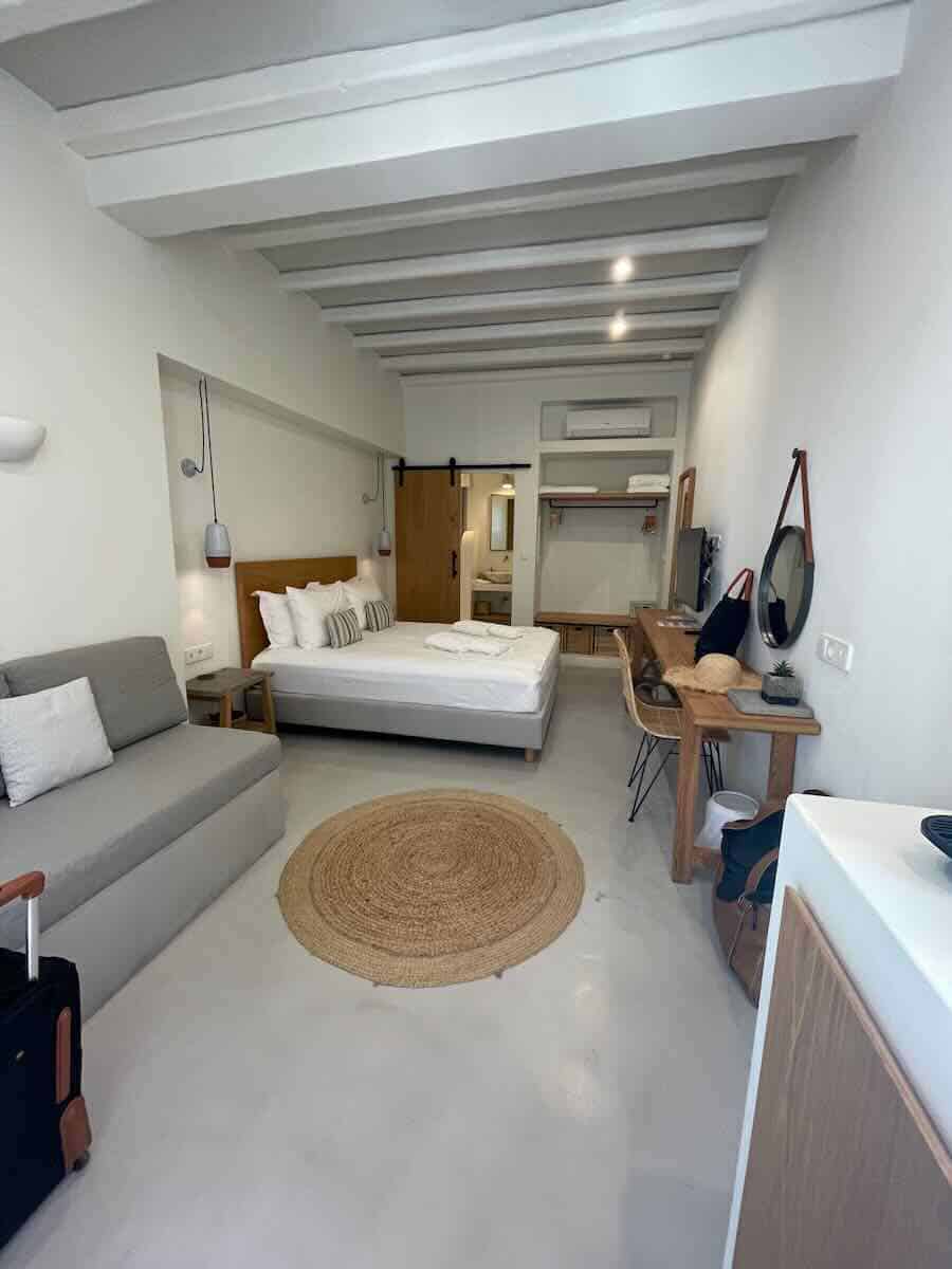 The image shows a modern, minimalist hotel room with a clean, open design. It features a bed, a gray sofa, a wooden desk, and a circular woven rug. The room has white walls with exposed beams and a sliding barn-style door leading to the bathroom. The space is cozy and functional, with simple yet stylish decor.