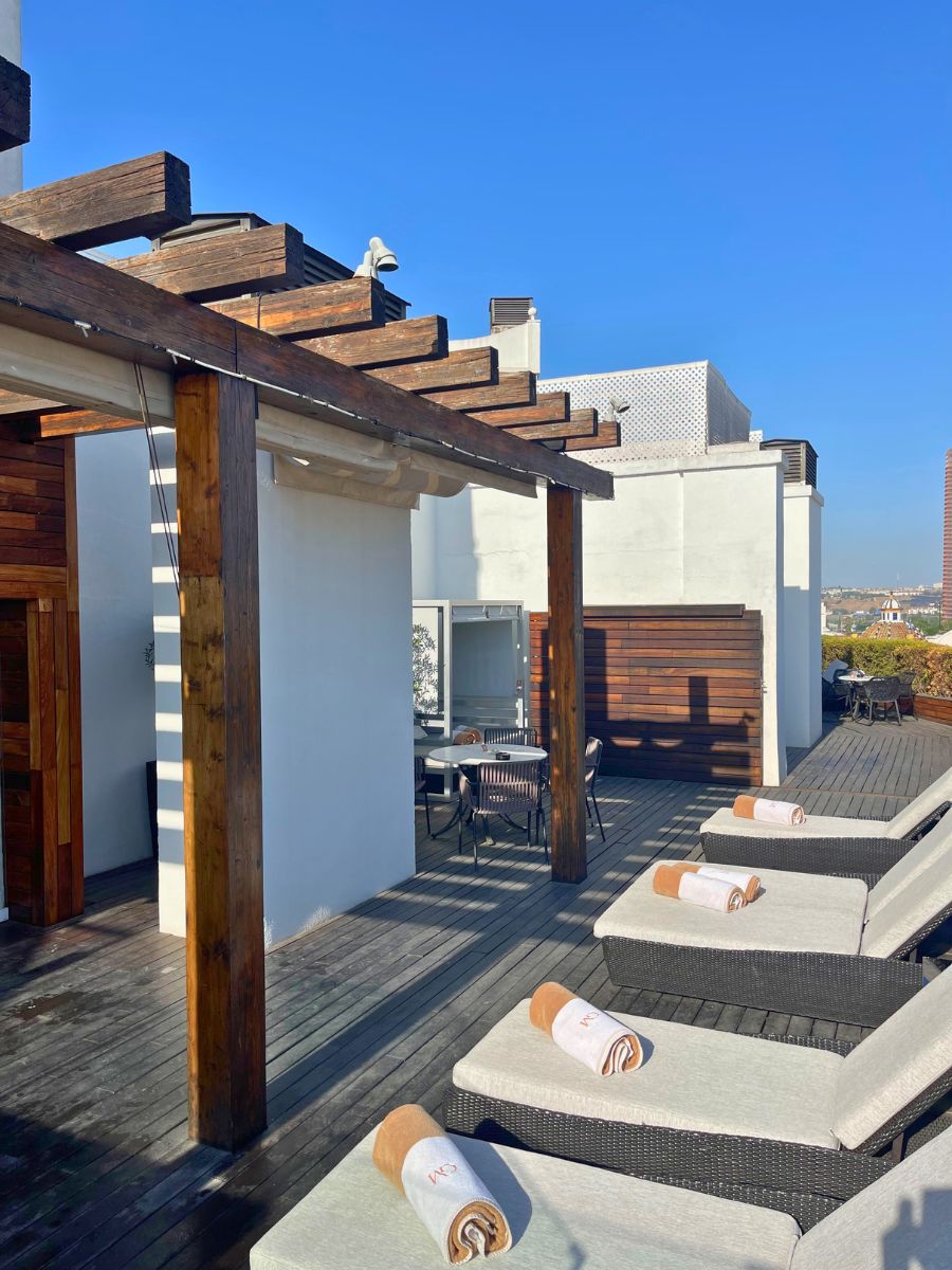 Lounge chairs with rolled towels under a wooden pergola on the rooftop terrace of a Seville hotel.