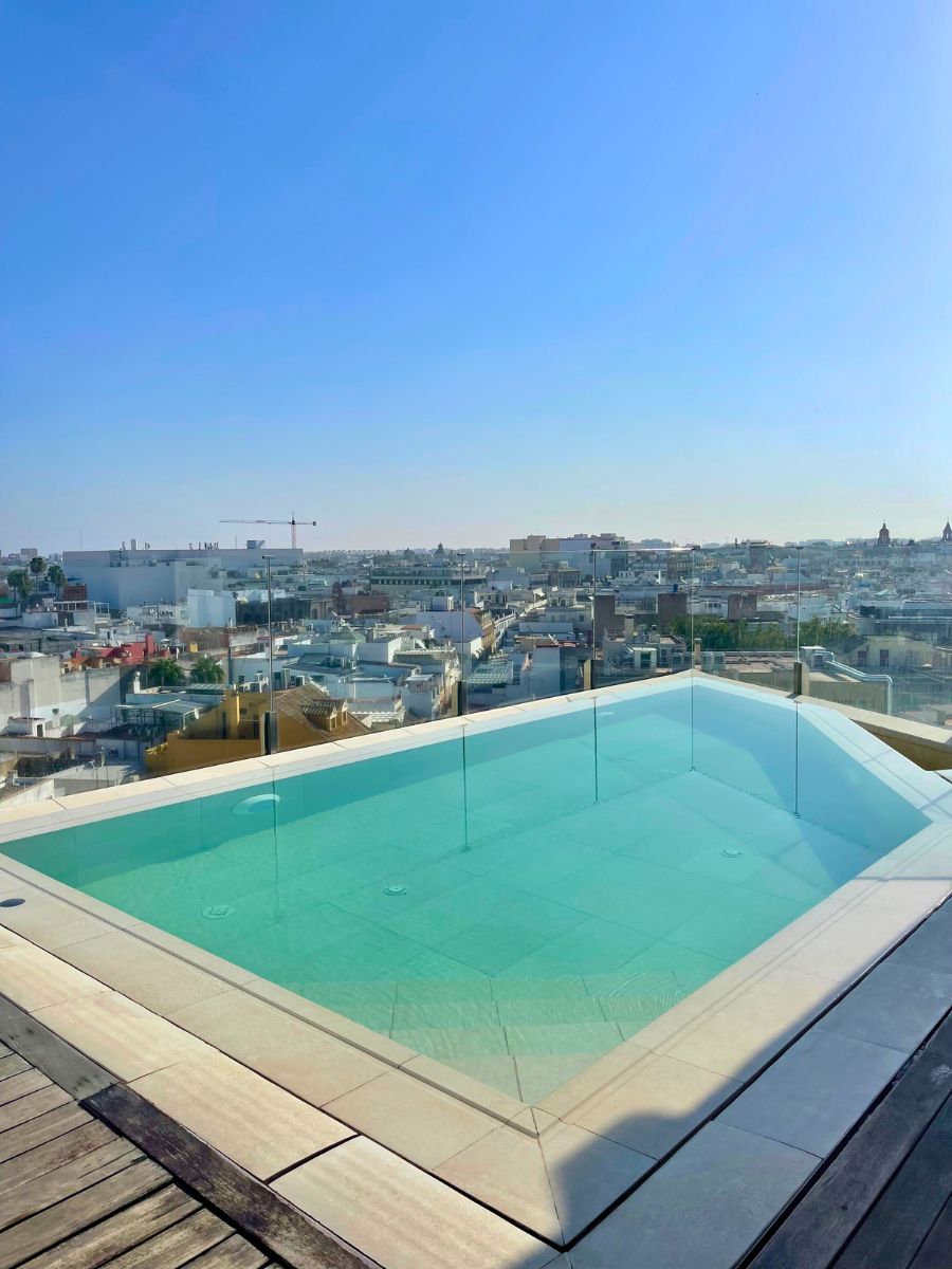 Small rooftop pool with glass panels overlooking Seville’s skyline under a clear blue sky.