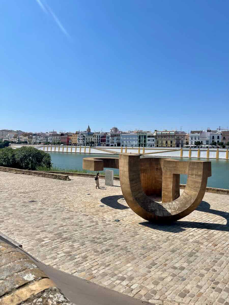 A large, modern sculpture located by the Guadalquivir River in Seville, Spain. The sculpture is abstract and geometric, with the river and a row of colorful buildings in the Triana neighborhood visible in the background under a clear blue sky.