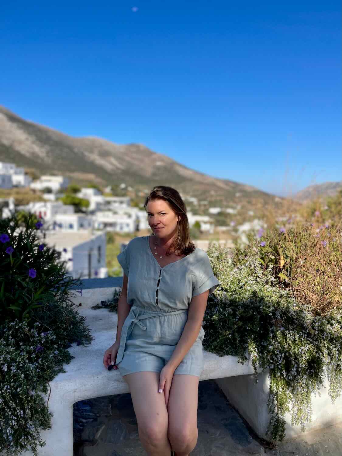 Woman in a casual outfit sitting on a white bench with a backdrop of lush greenery and white buildings in Sifnos.