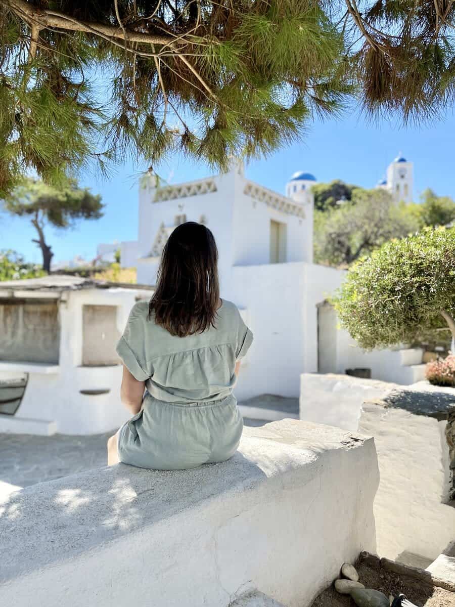 A woman sitting a ledge in Sifnos looking at the church in the background on a sunny day. 