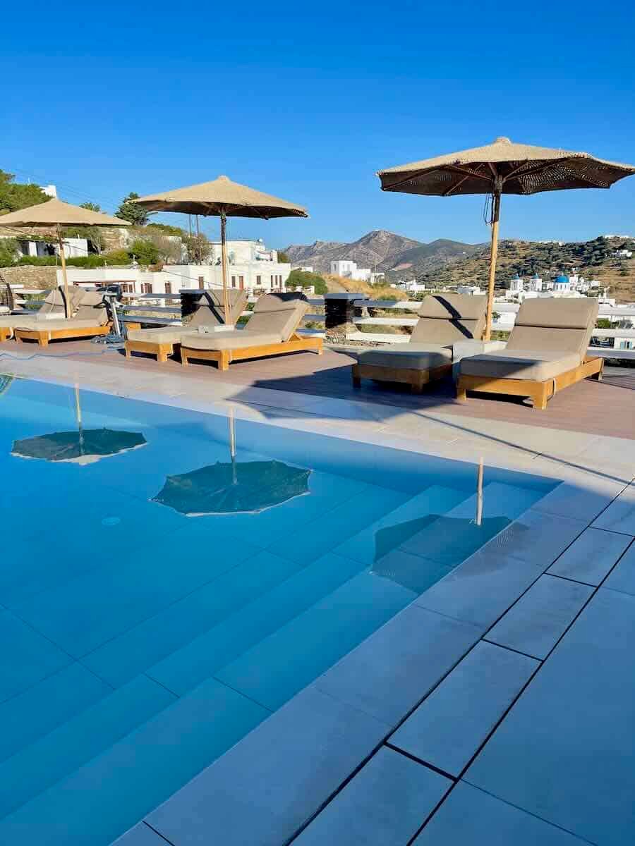 The image shows a luxurious poolside area with wooden lounge chairs and umbrellas set against a backdrop of clear blue skies and distant mountains. The calm, inviting pool reflects the umbrellas, and white-washed buildings are visible in the distance, suggesting a Mediterranean location. The scene is serene and perfect for relaxation.