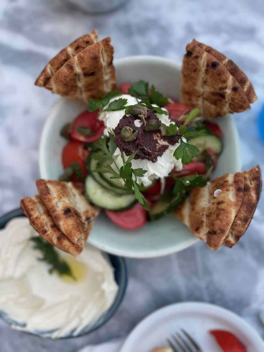 A fresh Greek salad with sliced cucumbers, cherry tomatoes, red onions, and feta cheese, garnished with olives and herbs. Grilled pita wedges are arranged around the bowl.