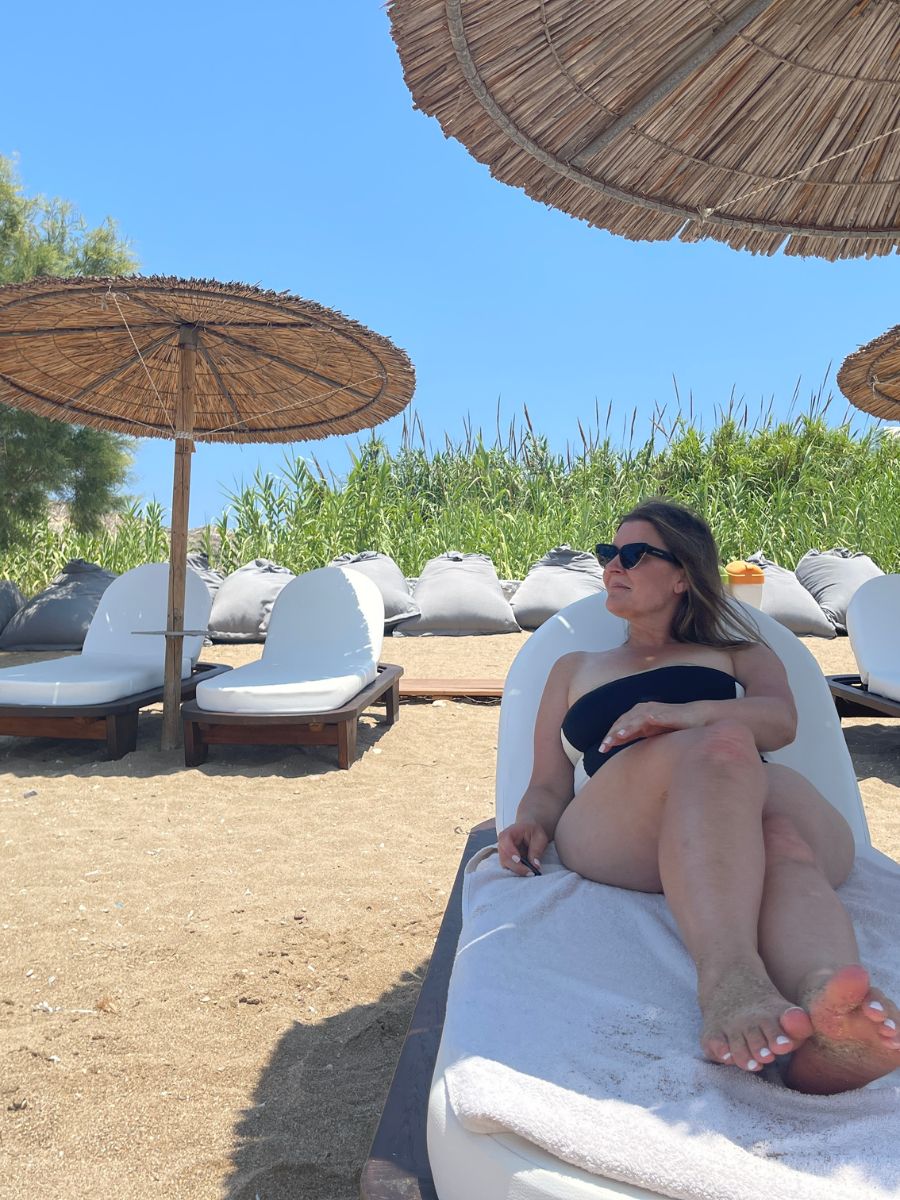 A woman in a black swimsuit lounging on a white beach chair under a straw umbrella on one of the serene beaches in Sifnos, Greece