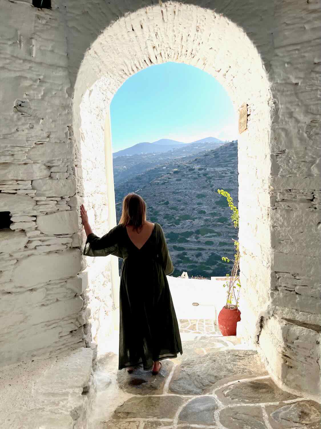 A person in a long dark dress stands in an arched stone doorway, looking out at a mountainous landscape under a clear sky. A potted plant adds a touch of greenery near the doorway, creating a serene, reflective scene.