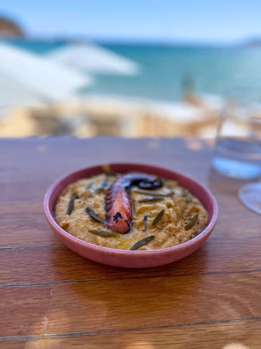 The image shows a bowl of food placed on a wooden table, with a scenic beach view in the background. The dish appears to be a creamy spread, possibly hummus or fava, topped with a grilled piece of octopus and garnished with herbs. The blurred background highlights the coastal setting, enhancing the relaxing atmosphere of dining by the sea.