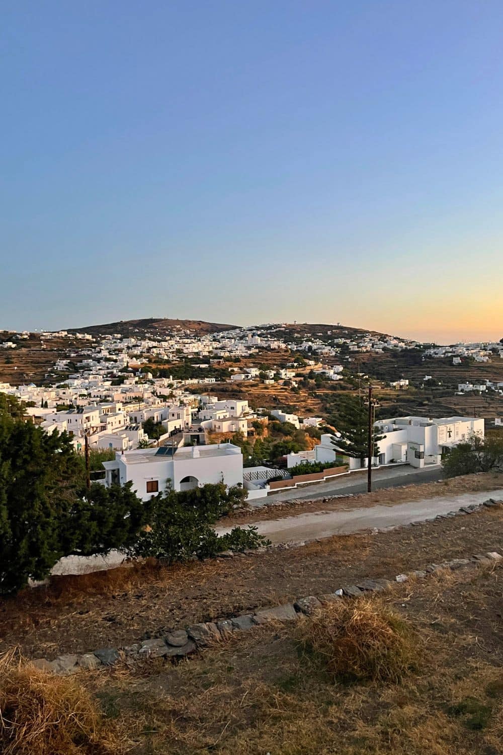 Twilight view of a sprawling Greek village with clustered white houses and rolling hills in the background.
