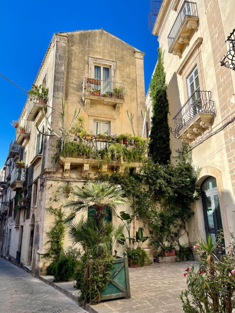 A charming historic building in Ortigia, Siracusa, Sicily, adorned with lush greenery and vibrant potted plants. The aged stone facade features small balconies with wrought-iron railings, decorated with cascading plants and flowers. Ivy climbs up the side of the adjacent building, adding to the old-world charm. A small courtyard entrance is framed by tropical plants, including a miniature palm tree. The narrow cobblestone street and warm sunlight enhance the Mediterranean atmosphere.