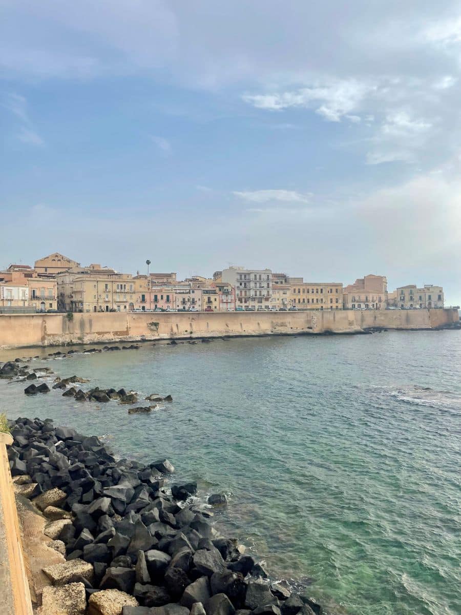 A scenic coastal view of Ortigia, the historic center of Siracusa, Sicily. The photo captures a row of pastel-colored buildings lined up behind an old stone seawall that stretches along the edge of the turquoise Mediterranean Sea. In the foreground, dark volcanic rocks contrast with the gentle waves lapping against the shore, while the sky above is lightly clouded, giving a soft and serene atmosphere.