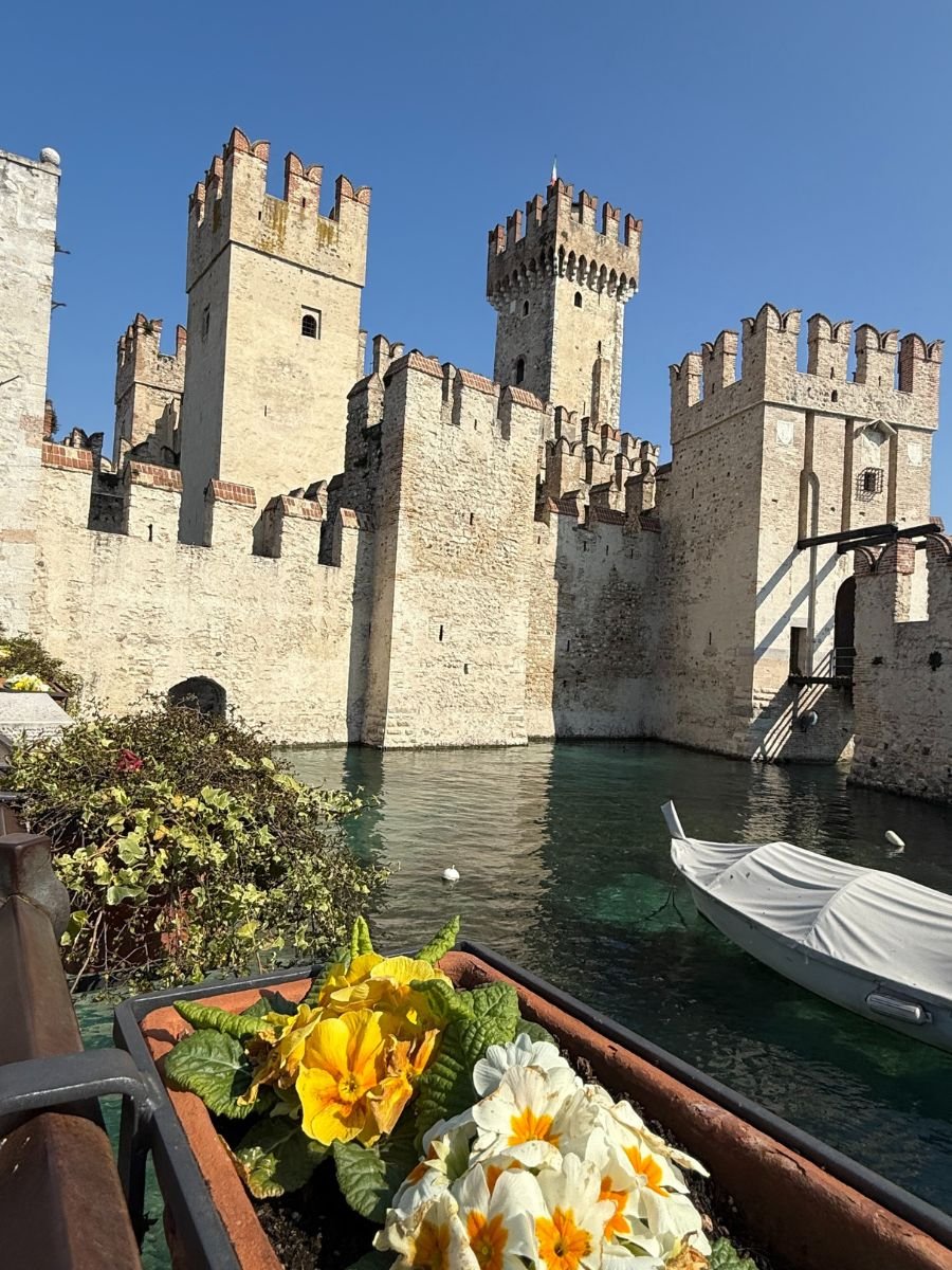 A picturesque view of Scaliger Castle in Sirmione, Italy, with its medieval stone walls and crenellated towers rising above the clear water of the moat. In the foreground, a planter box filled with bright yellow and white flowers adds a vibrant touch, while a covered boat floats gently on the water under a clear blue sky.