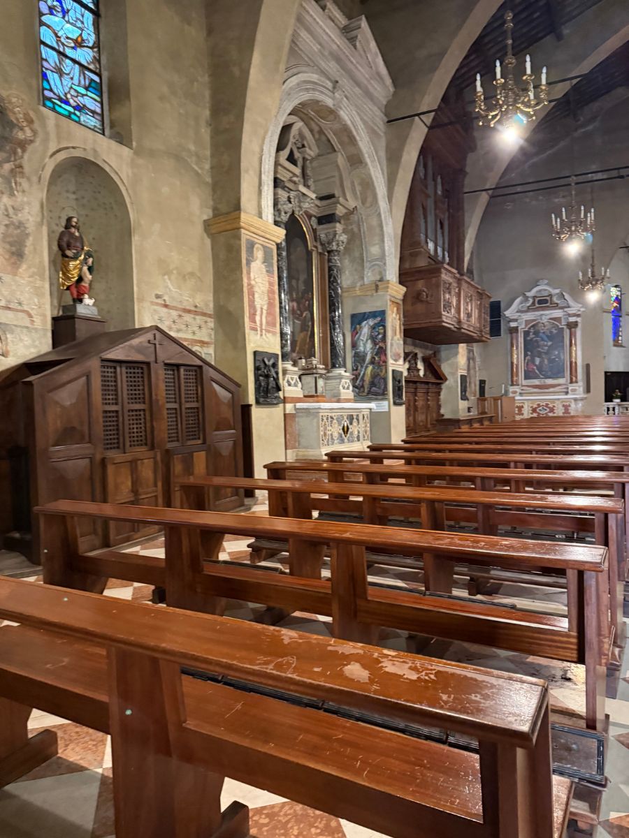 Interior of a historic church featuring wooden pews, arched stone architecture, and religious artwork. A confessional booth with a statue of a saint stands to the left, while altarpieces and paintings adorn the walls. Stained glass windows and chandeliers add to the solemn atmosphere.