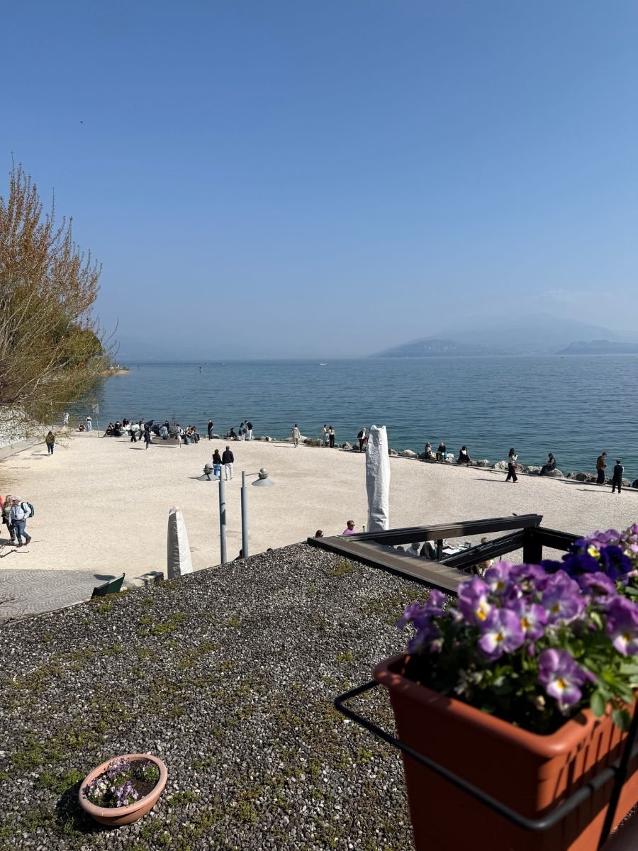 View overlooking a pebble beach on the shore of Lake Garda in Sirmione, Italy, with groups of people walking and relaxing near the water, flowering plants in pots in the foreground, and hazy mountains visible in the background under a clear blue sky.