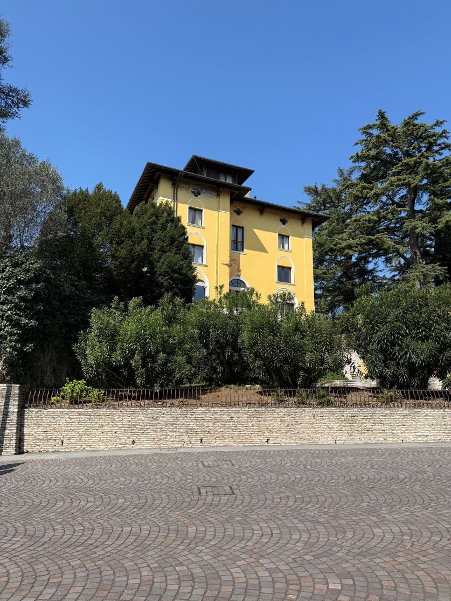 A bright yellow, four-story villa with arched windows, partially hidden behind lush green trees and shrubs, set against a clear blue sky in Sirmione, Italy. The foreground features a patterned cobblestone street and a low brick wall with an iron fence.