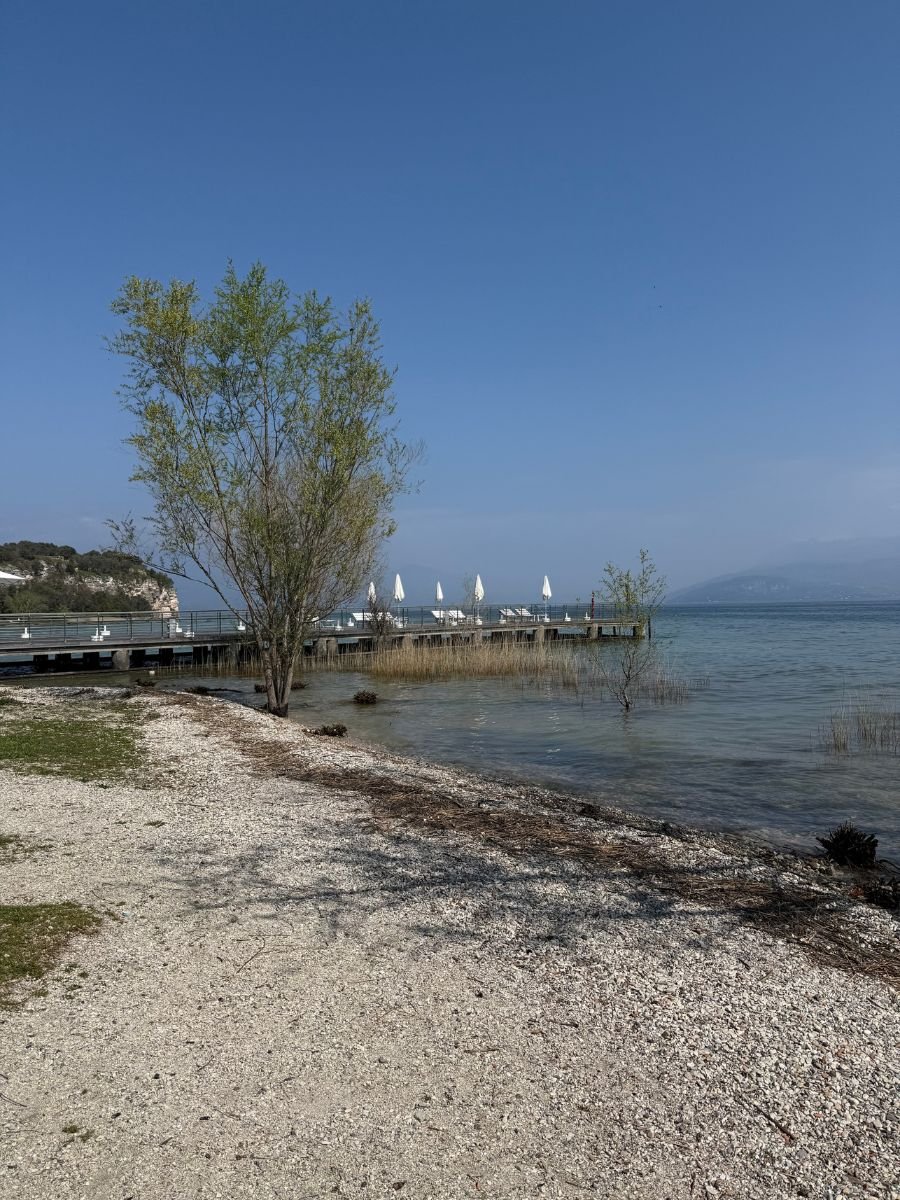 A peaceful lakeside scene in Sirmione, Italy, featuring a tree-lined shoreline with a gravel beach, a wooden pier extending into the clear blue water, and white closed umbrellas on the pier under a bright blue sky. Distant hills and light haze add depth to the tranquil landscape.