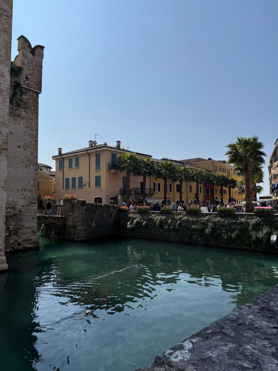 A canal with crystal-clear turquoise water flows beside a stone wall and colorful buildings in the historic center of Sirmione, Italy. People stroll along the waterfront lined with palm trees and flower boxes, while the corner of a medieval stone tower rises on the left, adding a touch of history to the sunlit, picturesque scene.