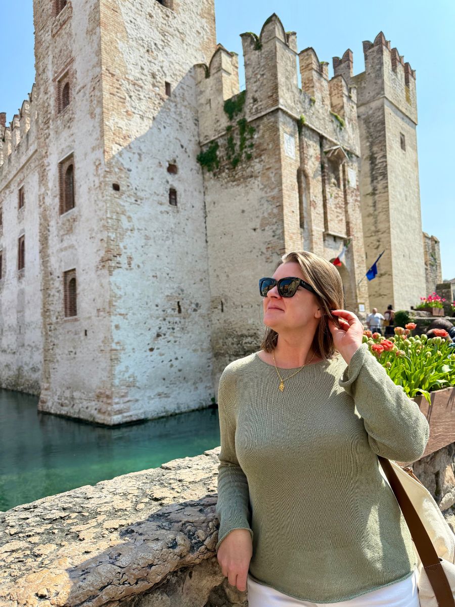 Melissa in a light green sweater and sunglasses stands by the stone wall of Scaliger Castle in Sirmione, Italy. With one hand touching her hair and the other resting on the ledge, she gazes upward. The medieval castle, with its crenellated towers and surrounding turquoise moat, rises behind her under a bright blue sky.