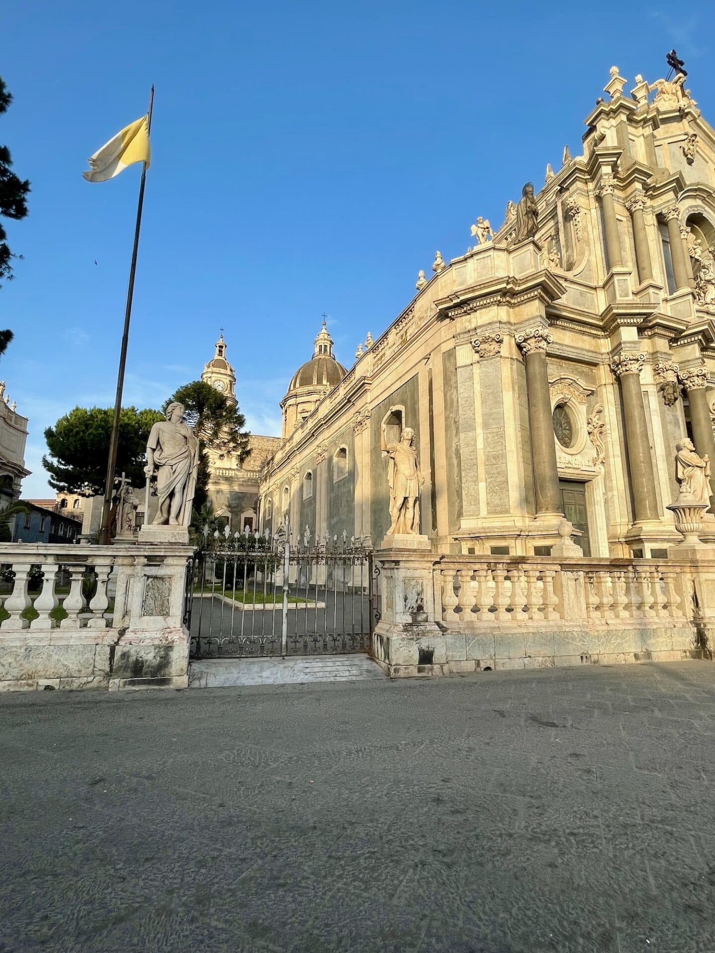 The exterior of a baroque church in Catania with statues and a flagpole in front.
