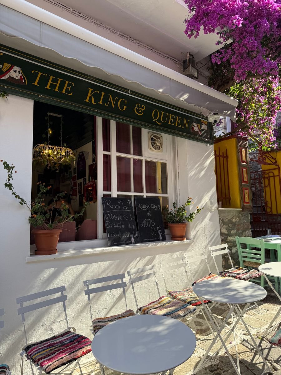 Outdoor seating of "The King & Queen" bar in Skiathos, Greece, with small round tables and white metal chairs topped with colorful woven cushions. A chalkboard menu advertises rooftop drinks, surrounded by potted plants and vibrant pink bougainvillea.