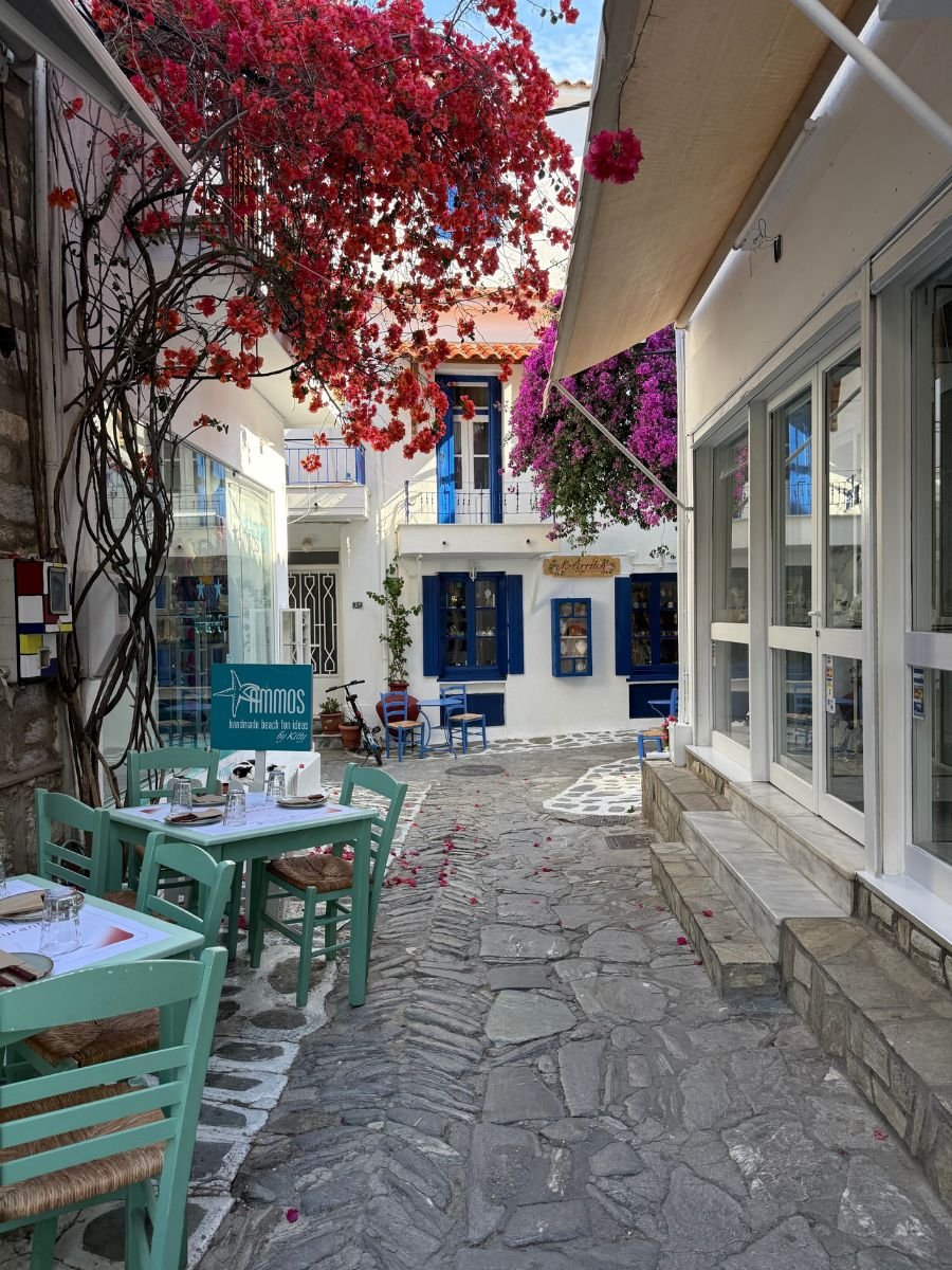 Charming cobblestone alley in Skiathos, Greece, lined with turquoise café tables and chairs, white buildings with blue shutters, and vibrant pink and purple bougainvillea flowers overhead.
