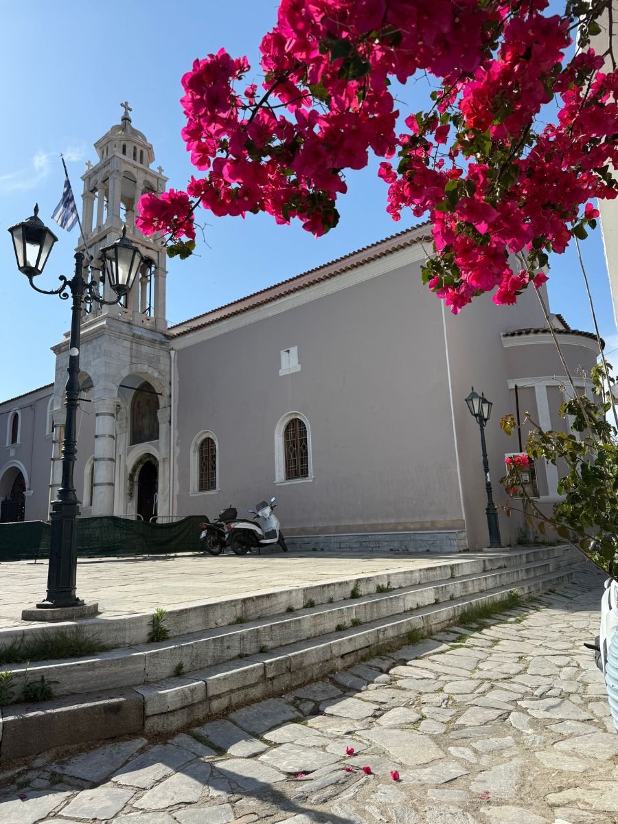 Historic Greek Orthodox church in Skiathos with a bell tower, stone steps, and bright pink bougainvillea flowers in the foreground under a clear blue sky.