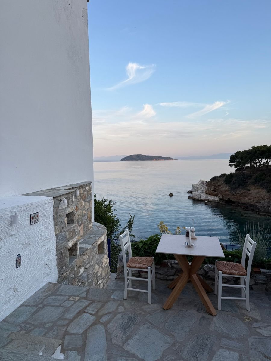 A quaint table for two set on a stone terrace overlooking the calm Aegean Sea at sunset, with an island and soft clouds in the background.