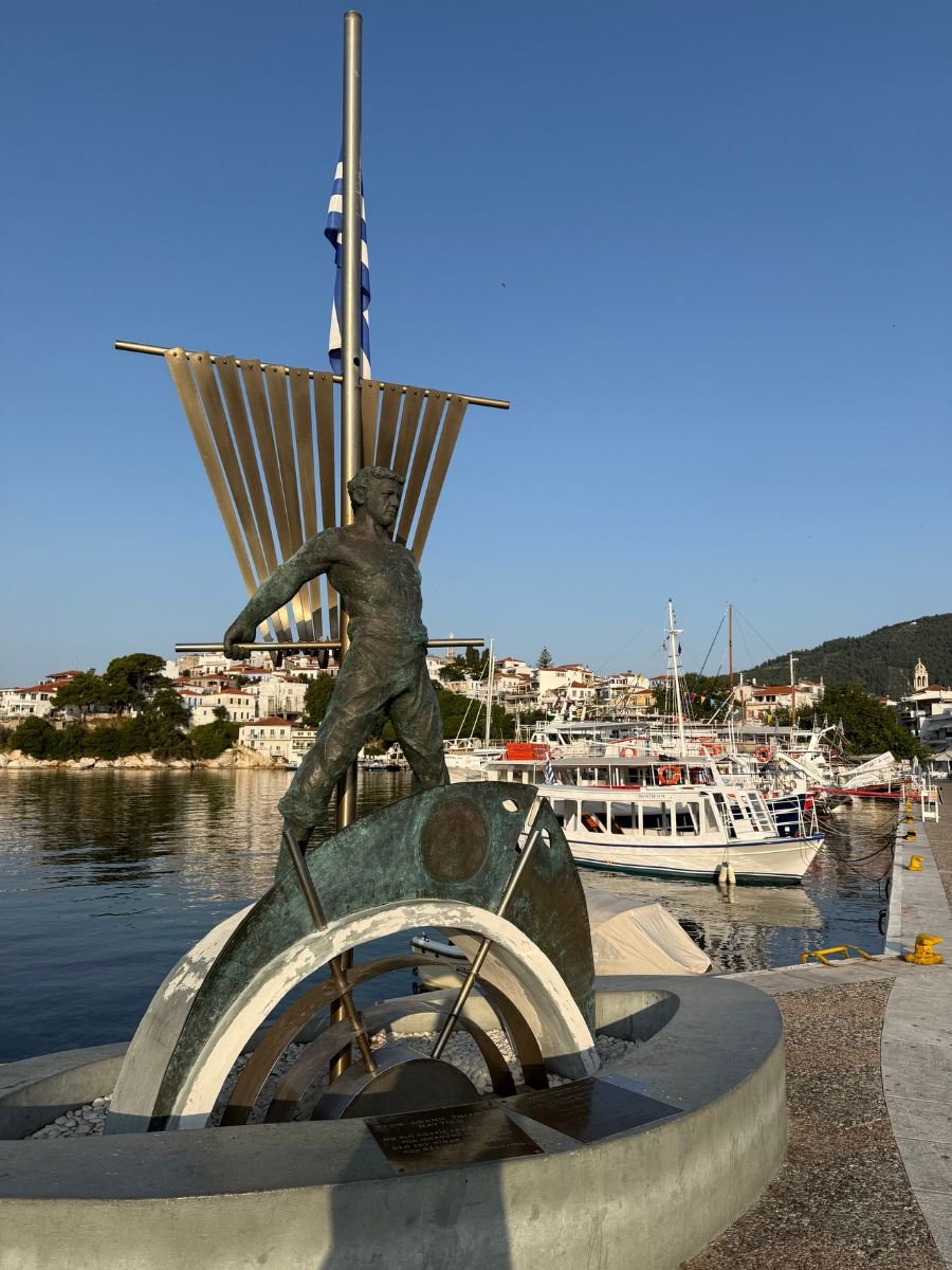 Bronze statue of a sailor standing on a stylized ship sculpture in Skiathos harbor, with boats and white houses in the background under a clear blue sky.