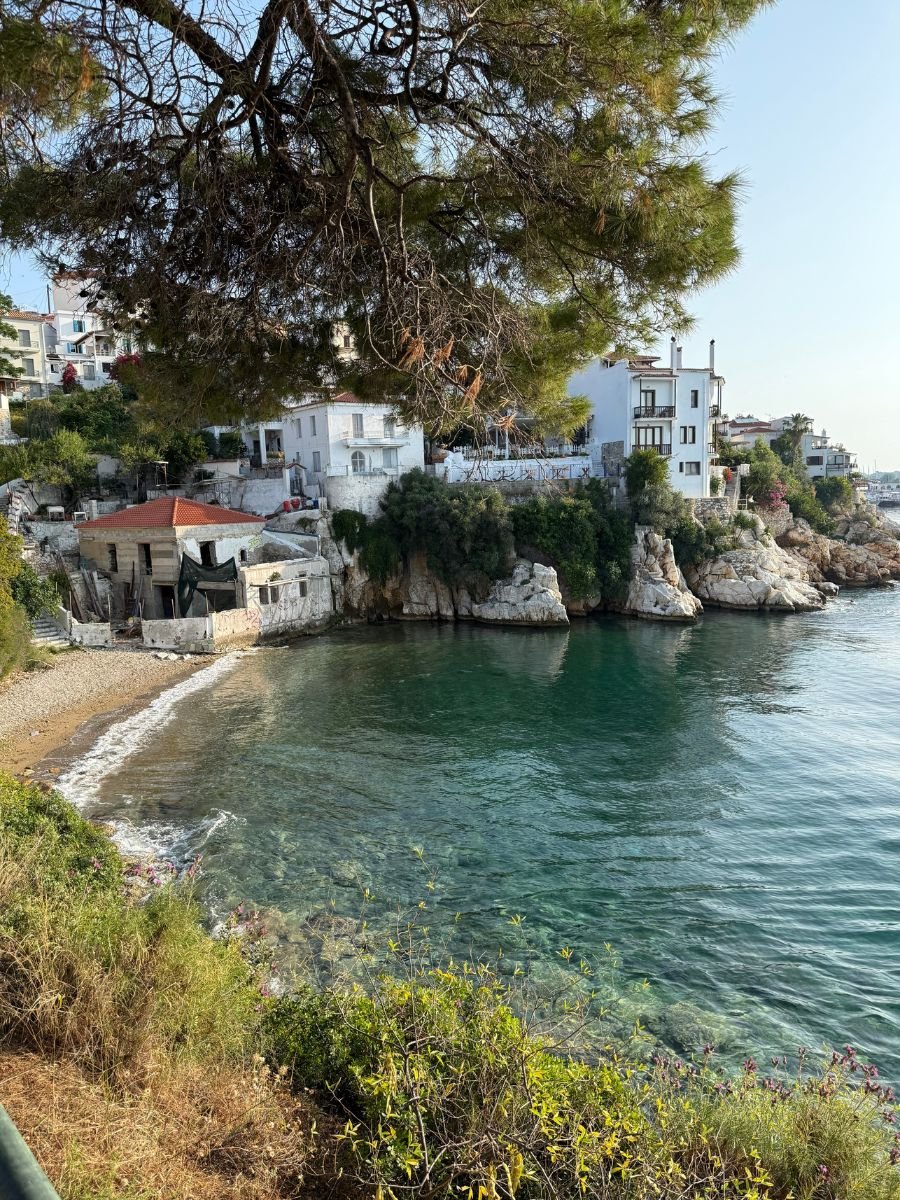 A peaceful coastal view in Skiathos featuring crystal-clear turquoise water, rocky cliffs with white-washed buildings perched above, and a small pebbled beach bordered by greenery.