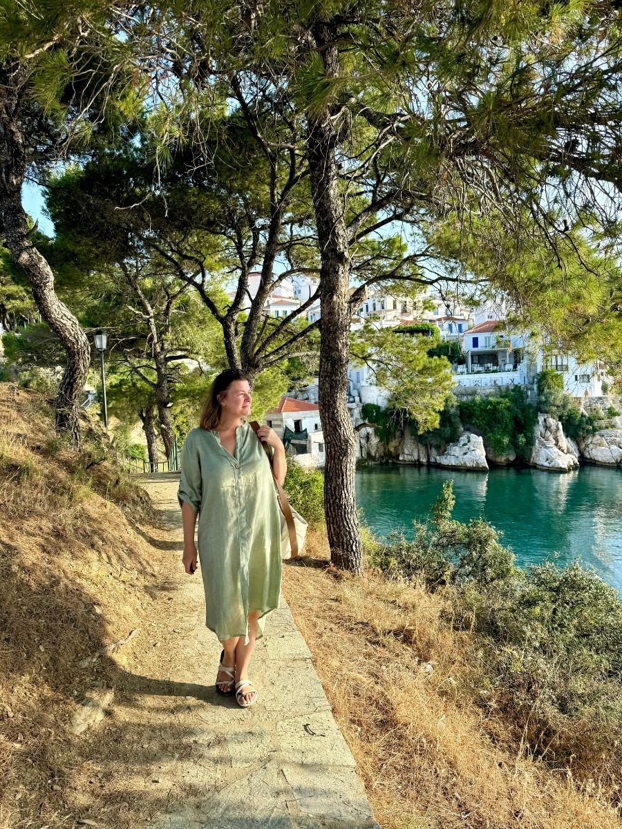 Melissa in a light green dress walks along a scenic coastal path shaded by pine trees, with clear turquoise water and hillside homes of Skiathos visible in the background.