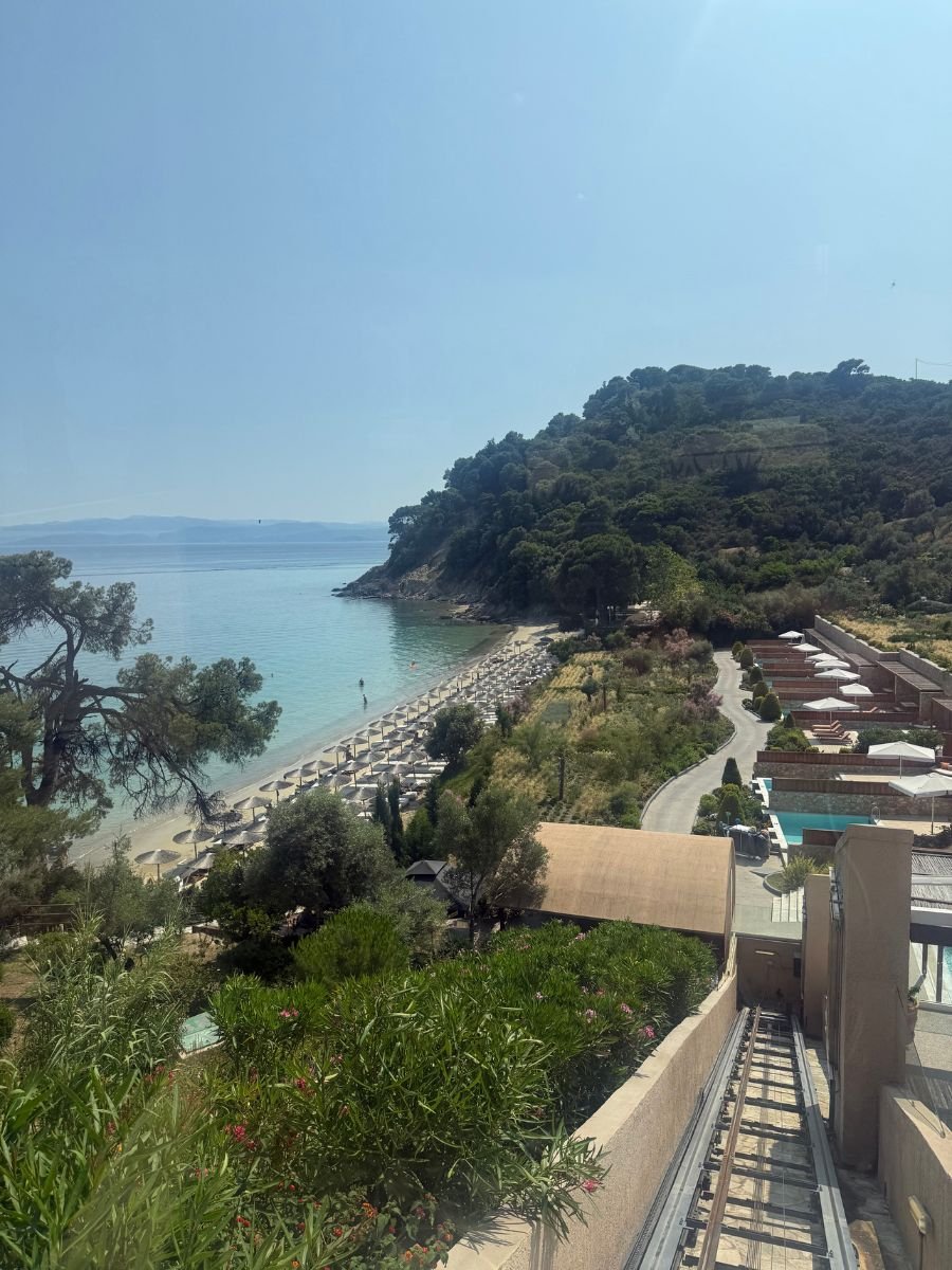 A panoramic view of a coastal resort in Skiathos, Greece, showing a funicular railway descending toward a beach lined with umbrellas and sunbeds, with lush greenery and a forested hill in the background.