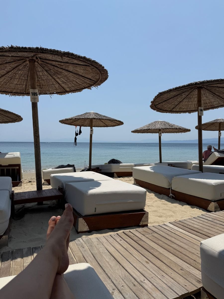 A relaxing view from a sunbed on a sandy beach in Skiathos, Greece, with rows of cushioned loungers and straw parasols facing the calm blue sea.