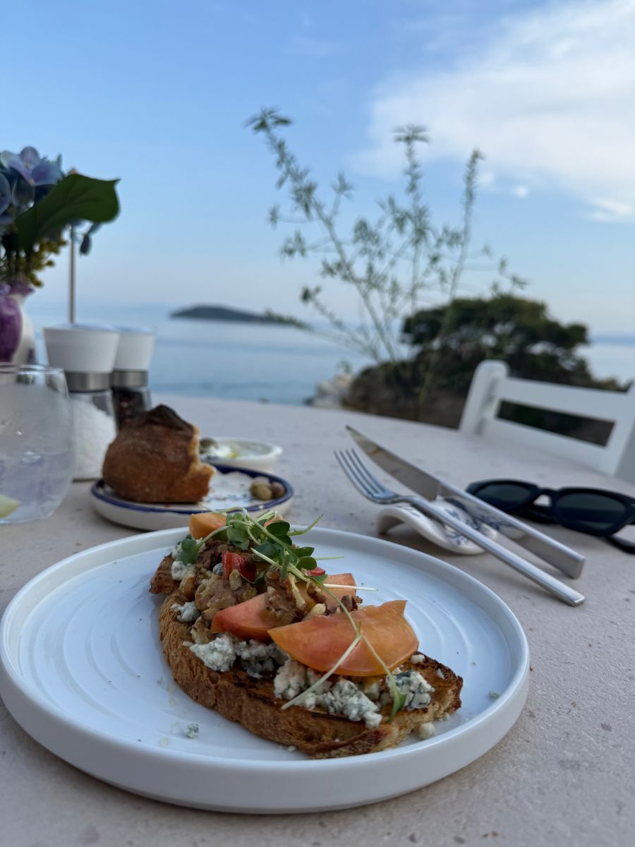 Gourmet open-faced sandwich topped with blue cheese, peaches, tomatoes, and microgreens on a white plate with a seaside view in the background.