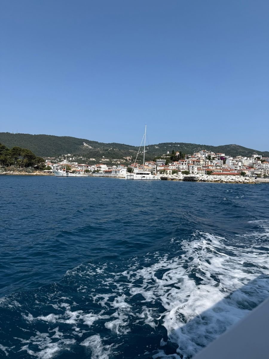 View from a boat approaching Skiathos Town, with deep blue Aegean waters in the foreground and white hillside buildings nestled against green mountains in the background.