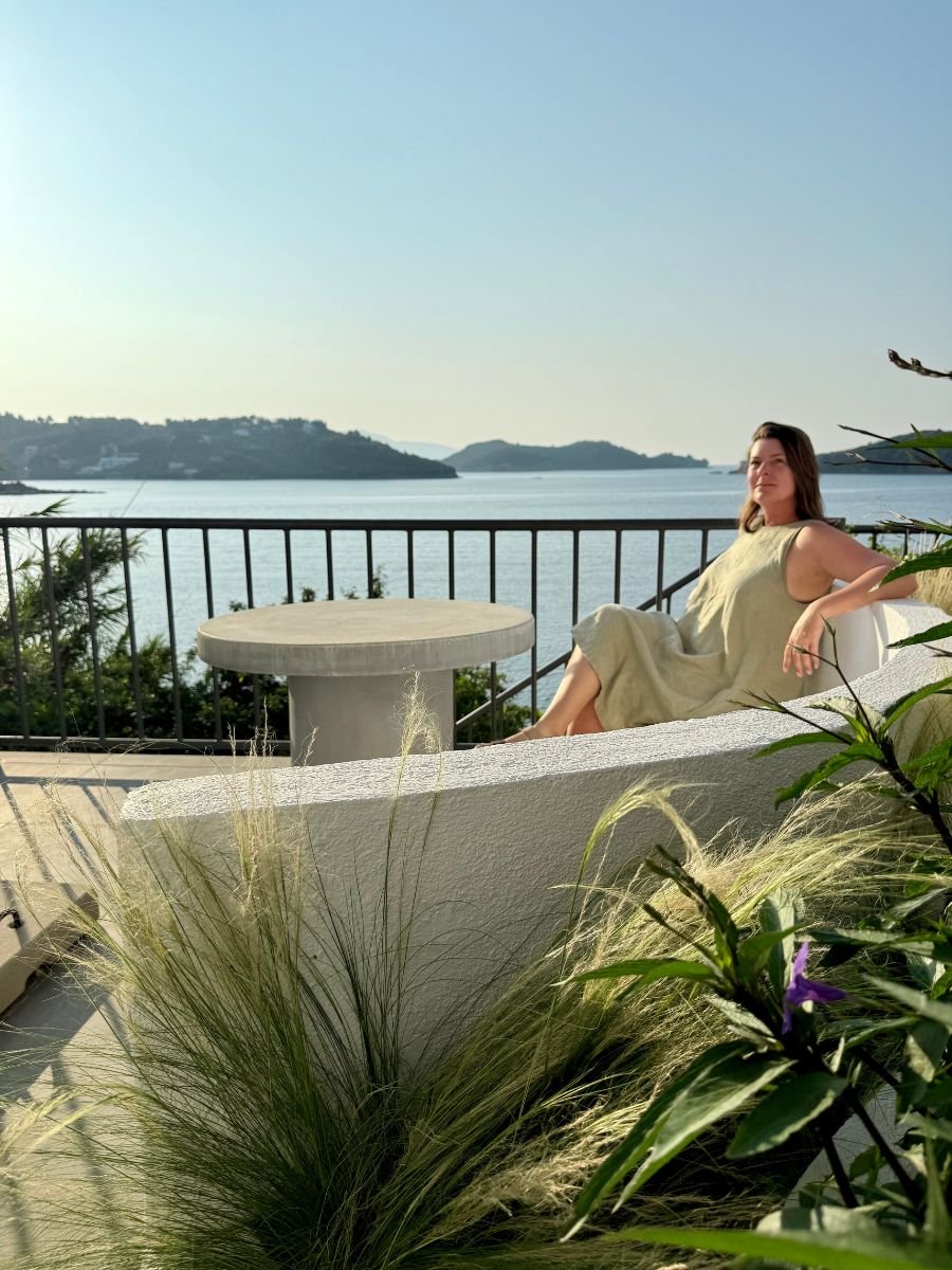 Melissa in a light green dress lounging on a curved white bench with a round table nearby, enjoying a serene view of the sea and distant green islands in Skiathos, surrounded by tall ornamental grasses and plants.