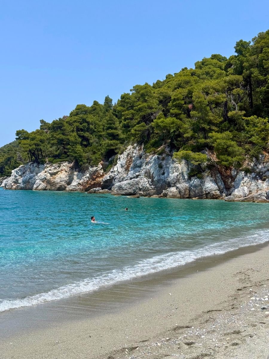 Peaceful beach scene with turquoise waters and swimmers near a rocky, tree-covered cliff on the island of Skopelos under a clear blue sky.