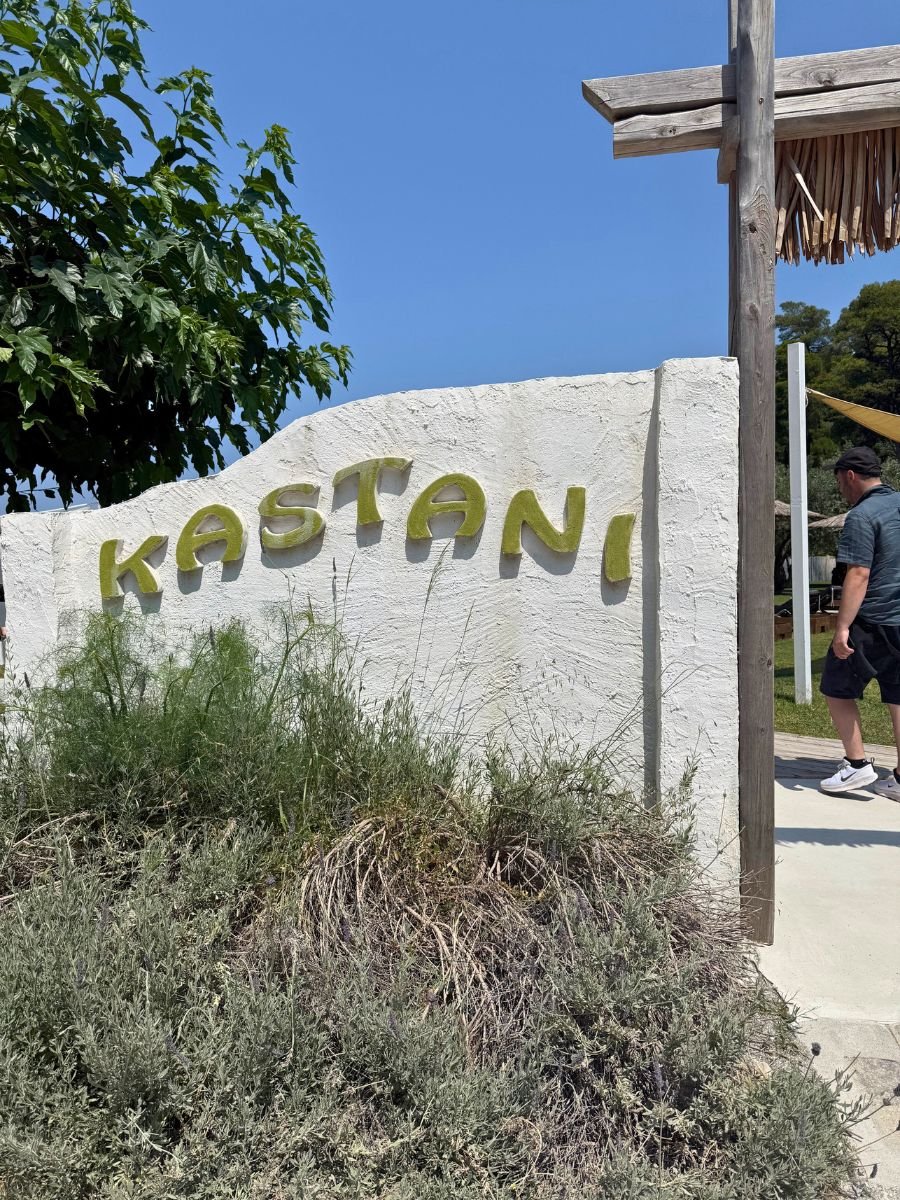 Entrance sign for Kastani Beach in Skopelos, Greece, with yellow lettering on a white wall surrounded by greenery.
