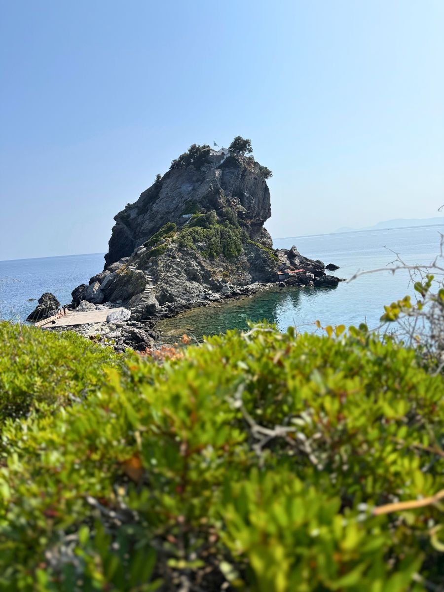 View of the iconic Agios Ioannis Chapel perched on a rocky hill above the Aegean Sea in Skopelos, with greenery in the foreground and clear blue skies above.