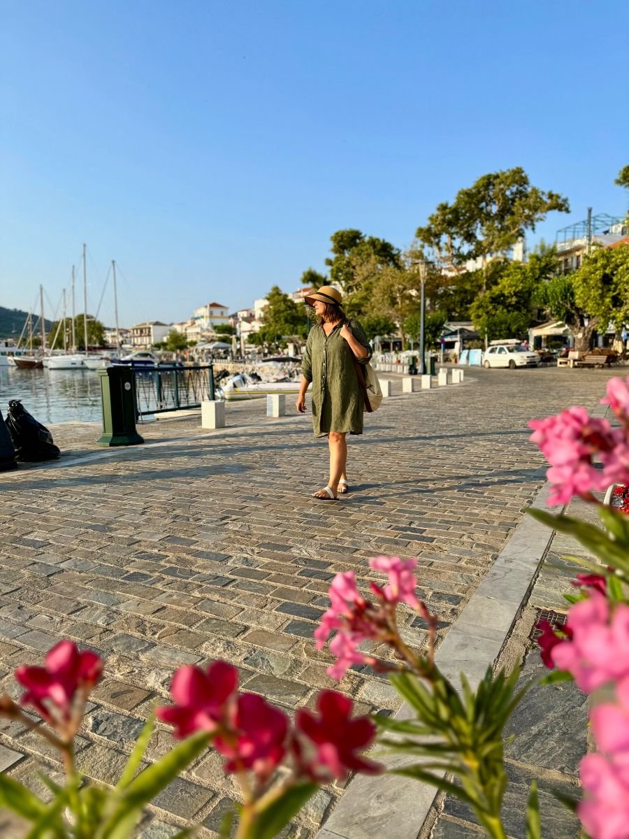 Melissa in a green dress and straw hat walking along a sunlit harbor promenade in Skopelos, Greece, with boats, flowers, and traditional buildings in the background.